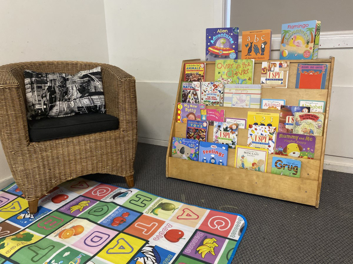A cosy reading corner with a wicker tub chair and cushions beside a picture-book display stand, set on a colourful alphabet play mat