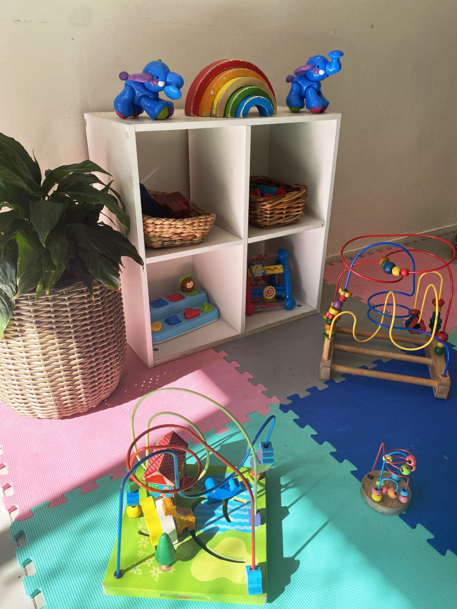 A toy shelf topped with a wooden rainbow stacker and two blue elephant push-toys, with bead-maze toys, baskets and a leafy pot plant on soft foam play mats