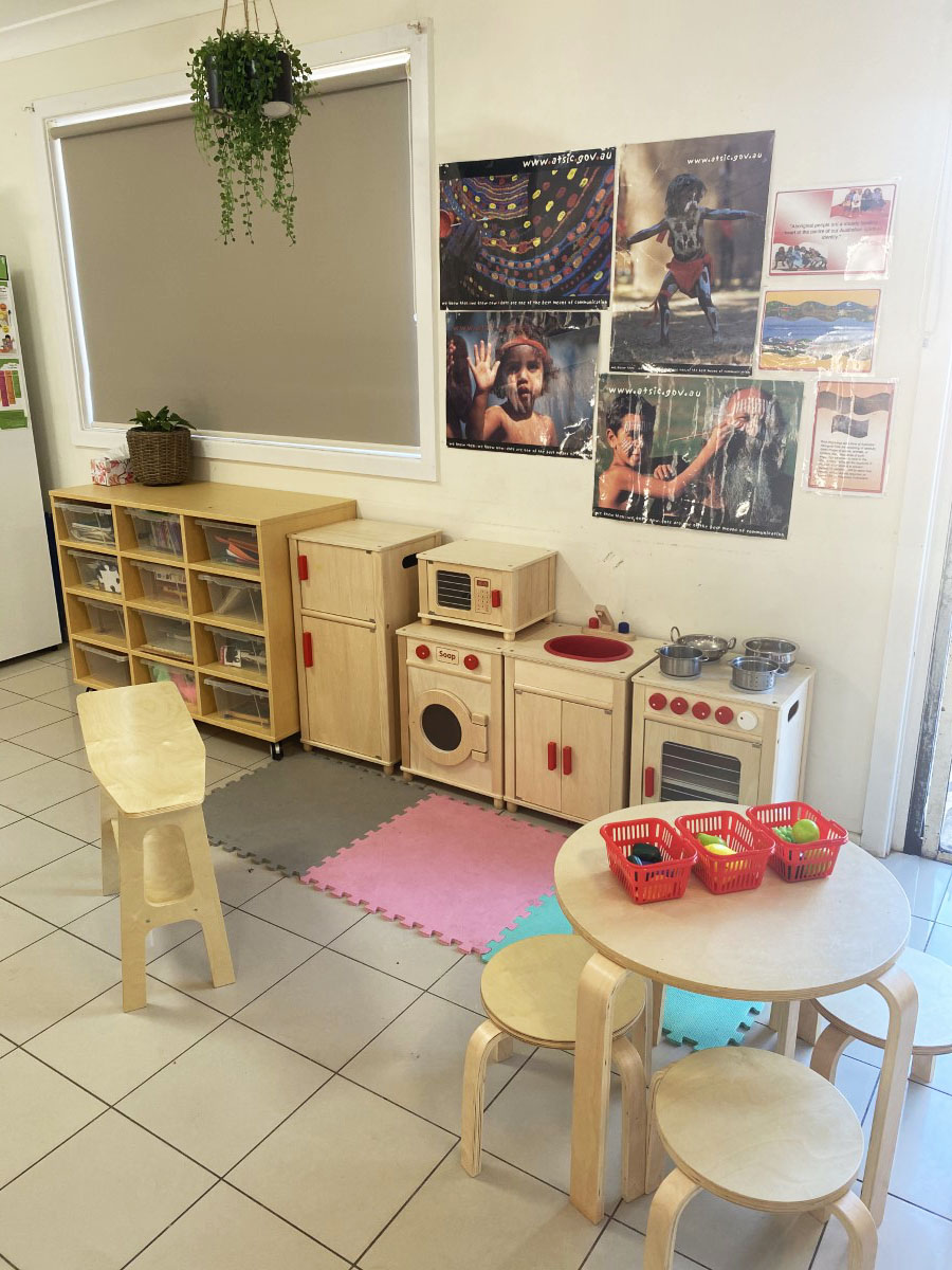 The home-corner play kitchen — a wooden fridge, washing machine, sink and oven with a round table of play-food baskets, below cultural posters on the wall