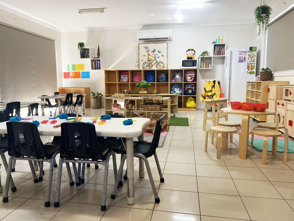 A bright, spacious classroom with low tables and little chairs, cube shelves of labelled bags and a home-corner play kitchen in natural light
