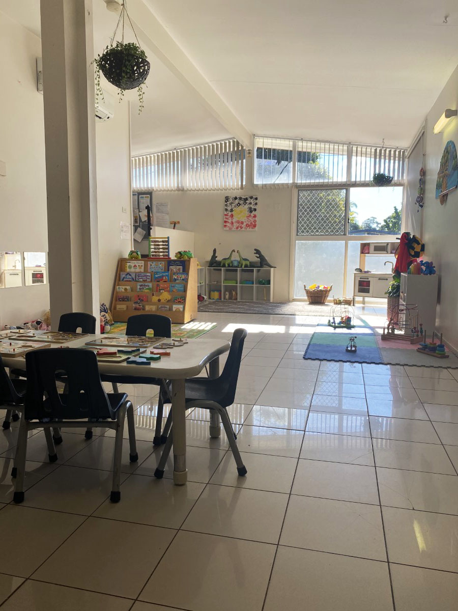 A light-filled playroom with a high vaulted ceiling and a wall of windows, a picture-book stand and a home-corner play kitchen on a tiled floor
