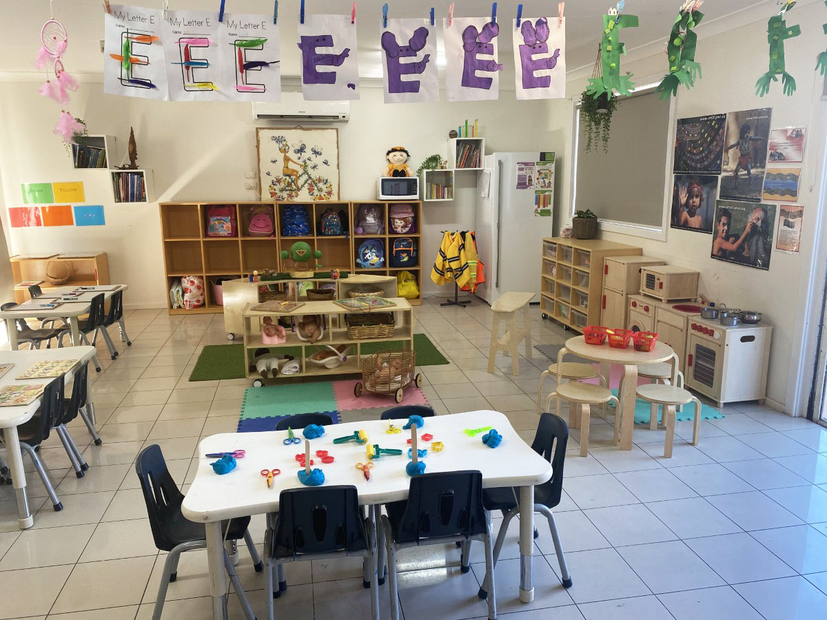 A wide view of a playroom with a central table of colourful play-dough, children’s letter artwork strung overhead and cube shelves of backpacks along the wall