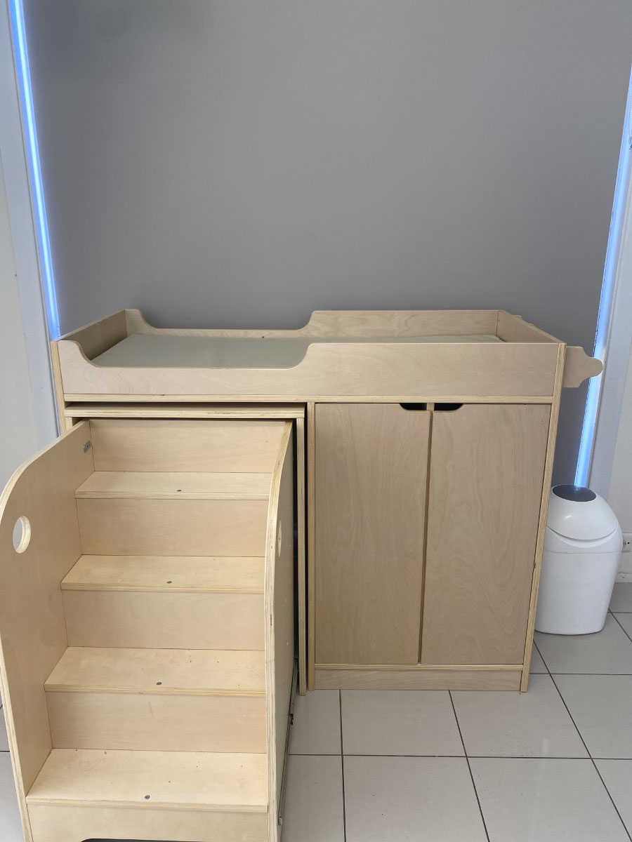 A wooden nappy-change station with built-in toddler steps and a storage cupboard beside a nappy bin, in a clean tiled room