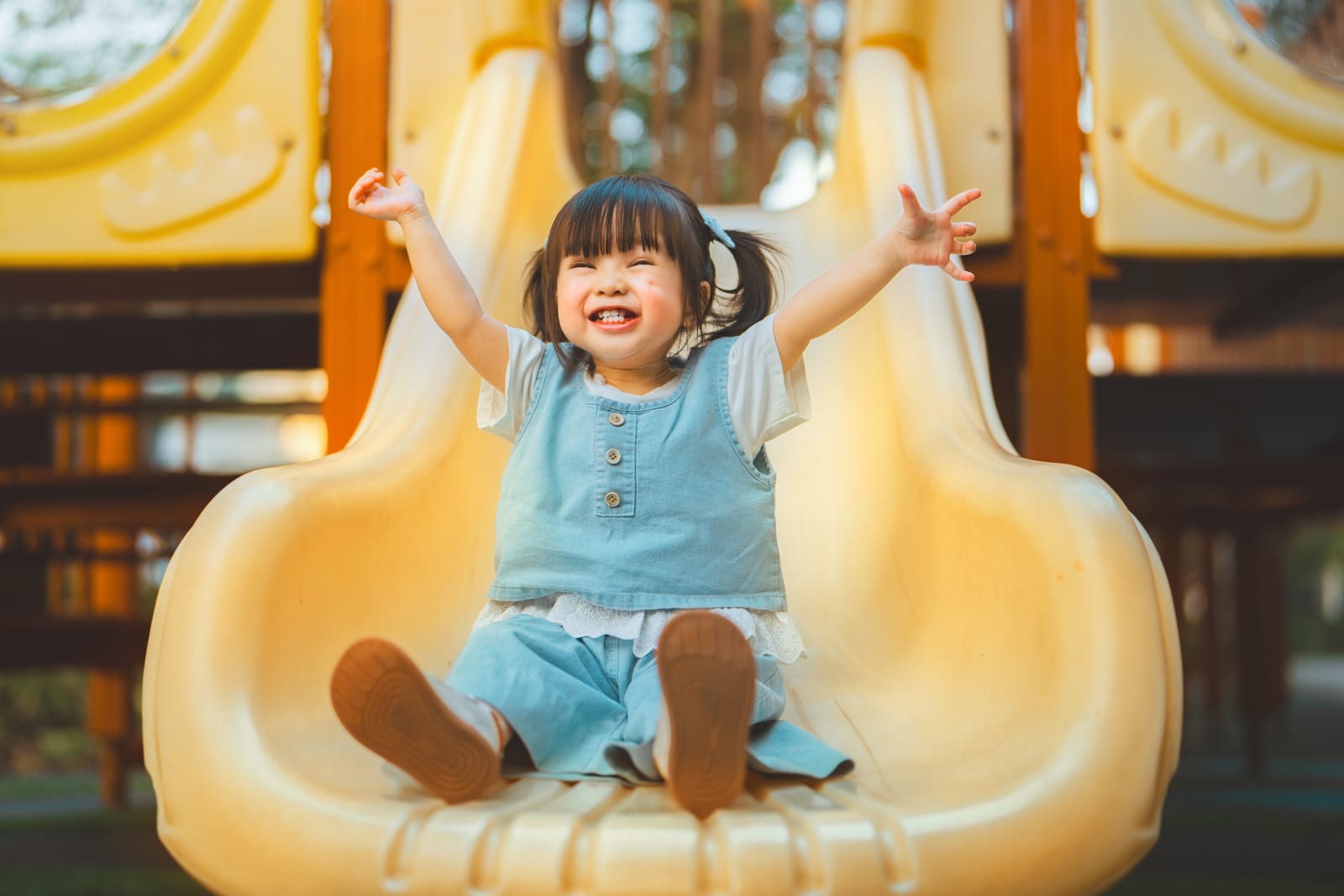 A happy toddler sitting at the bottom of a yellow playground slide with both arms raised in the air