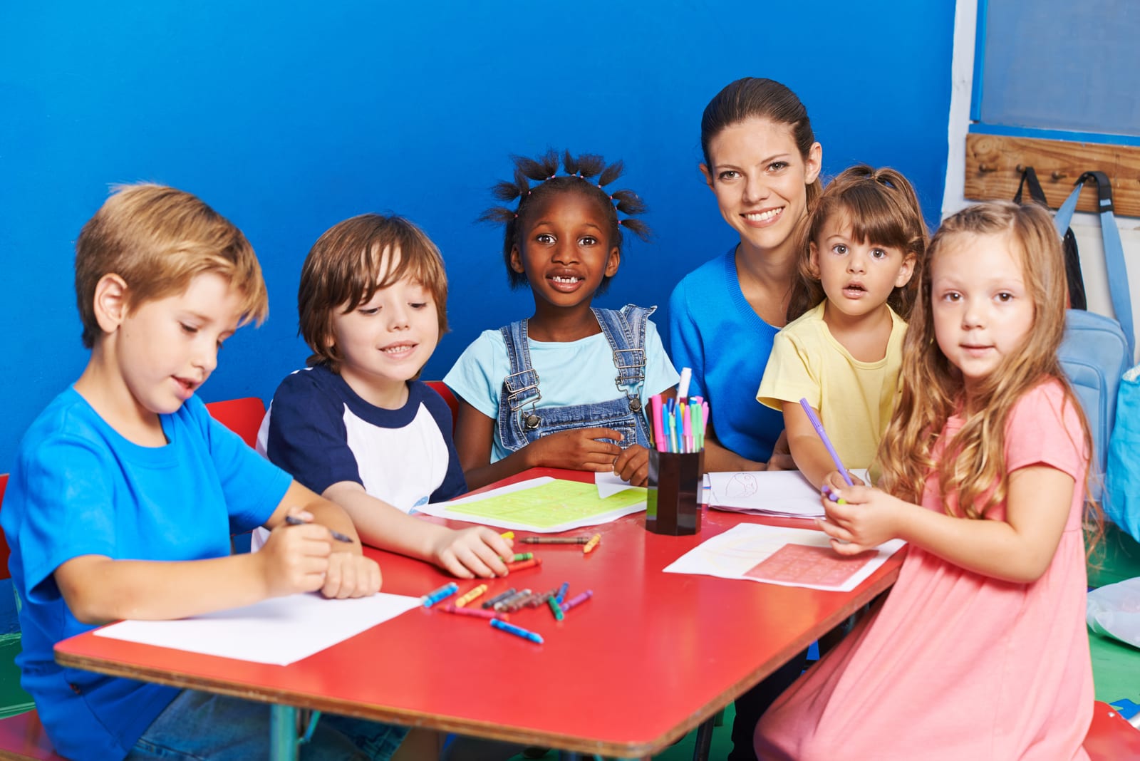 A smiling early-learning educator and a diverse group of young children drawing together at a red activity table