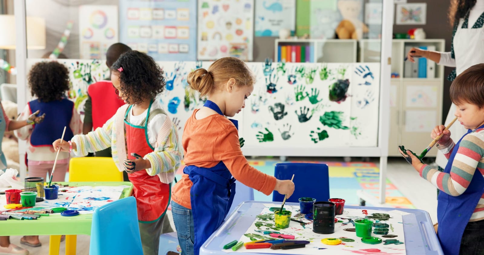 Young children in paint smocks painting at tables in a bright, colourful art room, with their handprint artwork covering the cabinet behind them