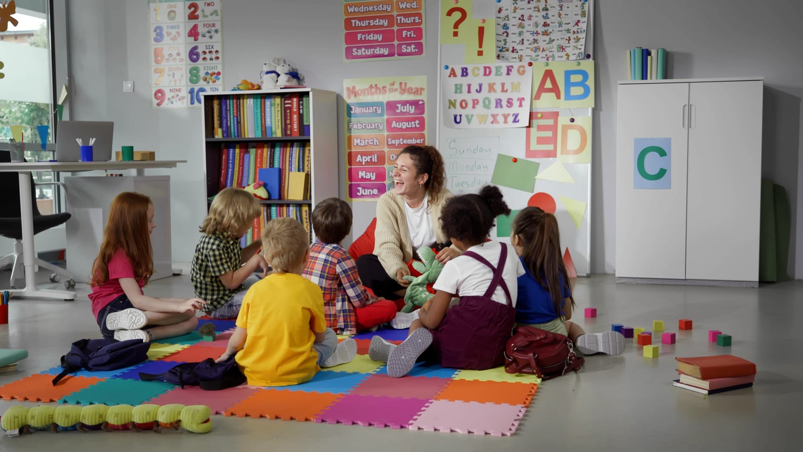 An educator sitting on a colourful foam play mat with a group of young children in a classroom lined with alphabet and number posters