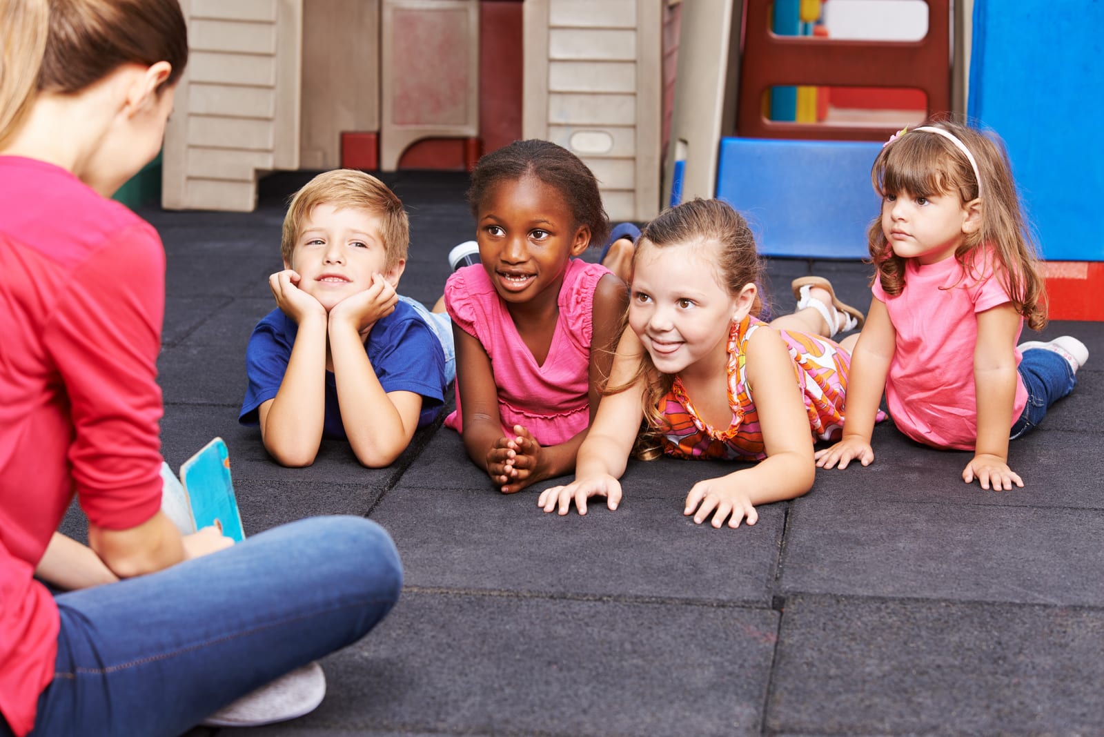 An educator reading to four young children who lie on their tummies on a soft play mat, listening