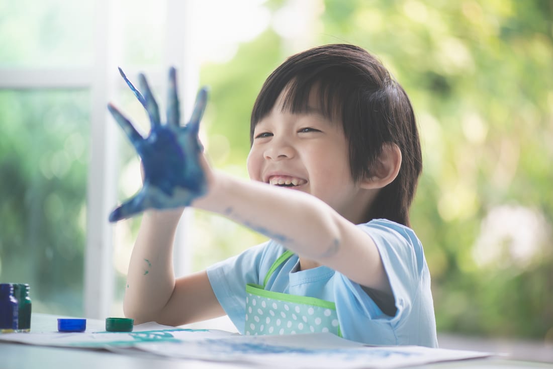 A smiling young child proudly showing a hand covered in blue paint during an art activity