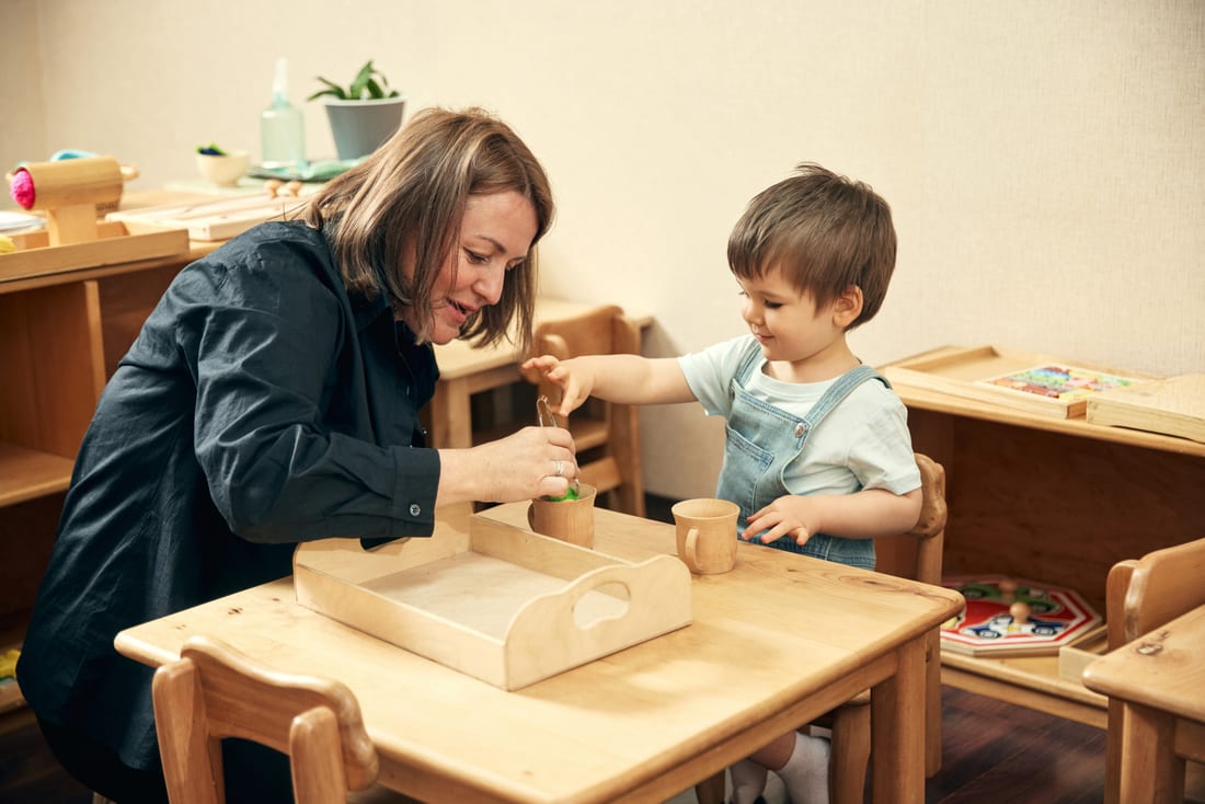 An educator and a toddler exploring a hands-on wooden pouring activity together at a low table